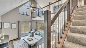 Un salón amplio con techo alto, una escalera con barandillas de madera y vistas al exterior a través de ventanas grandes.