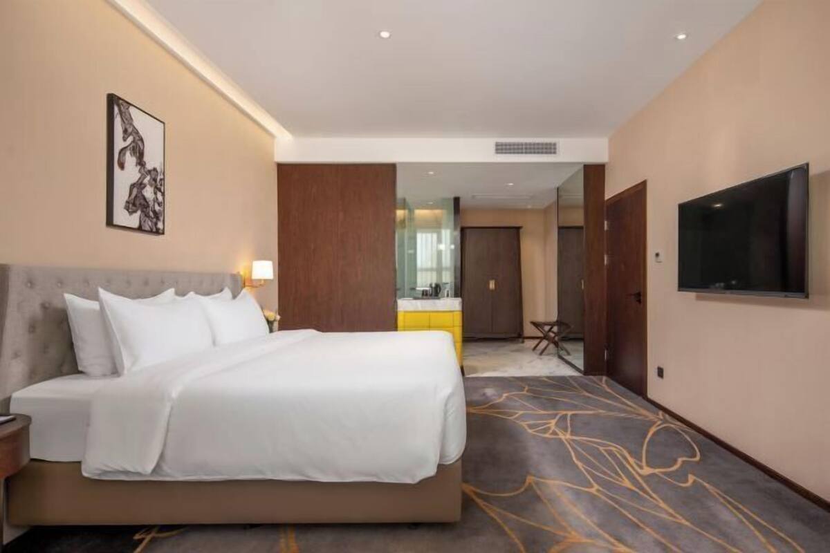 A hotel room with a bed, a TV, a painting, and a bathroom visible through a glass door.
