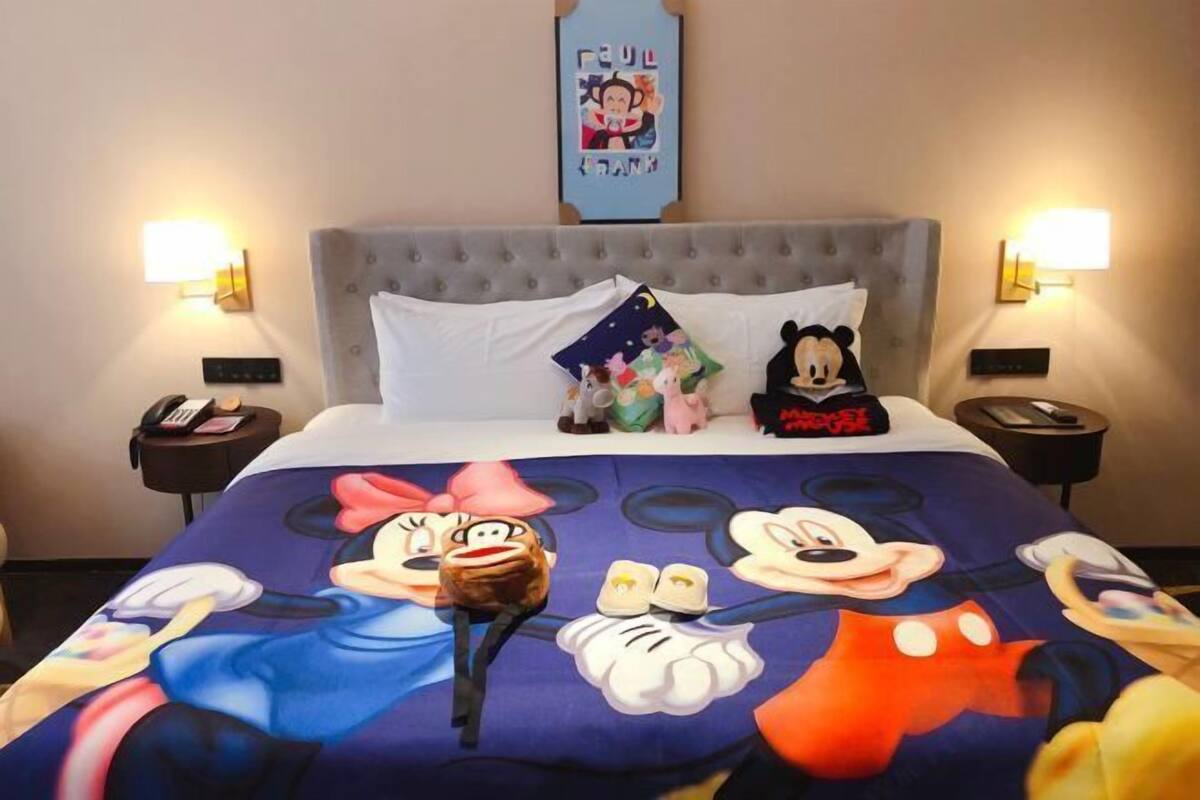 A hotel room with a bed featuring a Mickey Mouse themed bedspread, a tufted headboard, two bedside tables with lamps, and a wall-mounted poster.