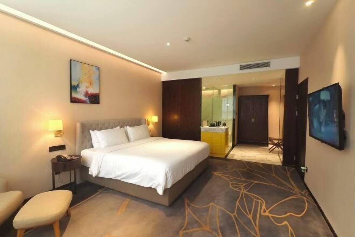 A hotel room with a bed, bedside table, chair, artwork, and a bathroom visible through an open door.