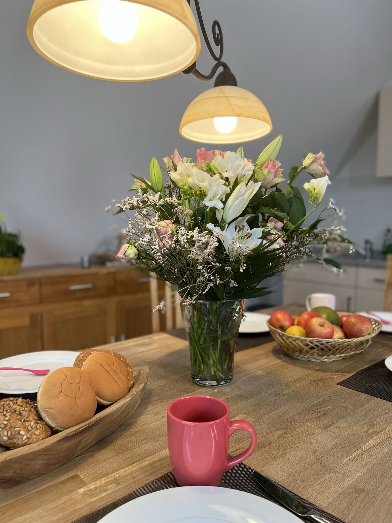 A dining table with a vase of flowers, a basket of apples, and a pink mug.