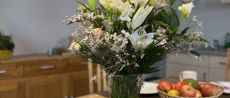 A dining table with a vase of flowers, a basket of apples, and a pink mug.