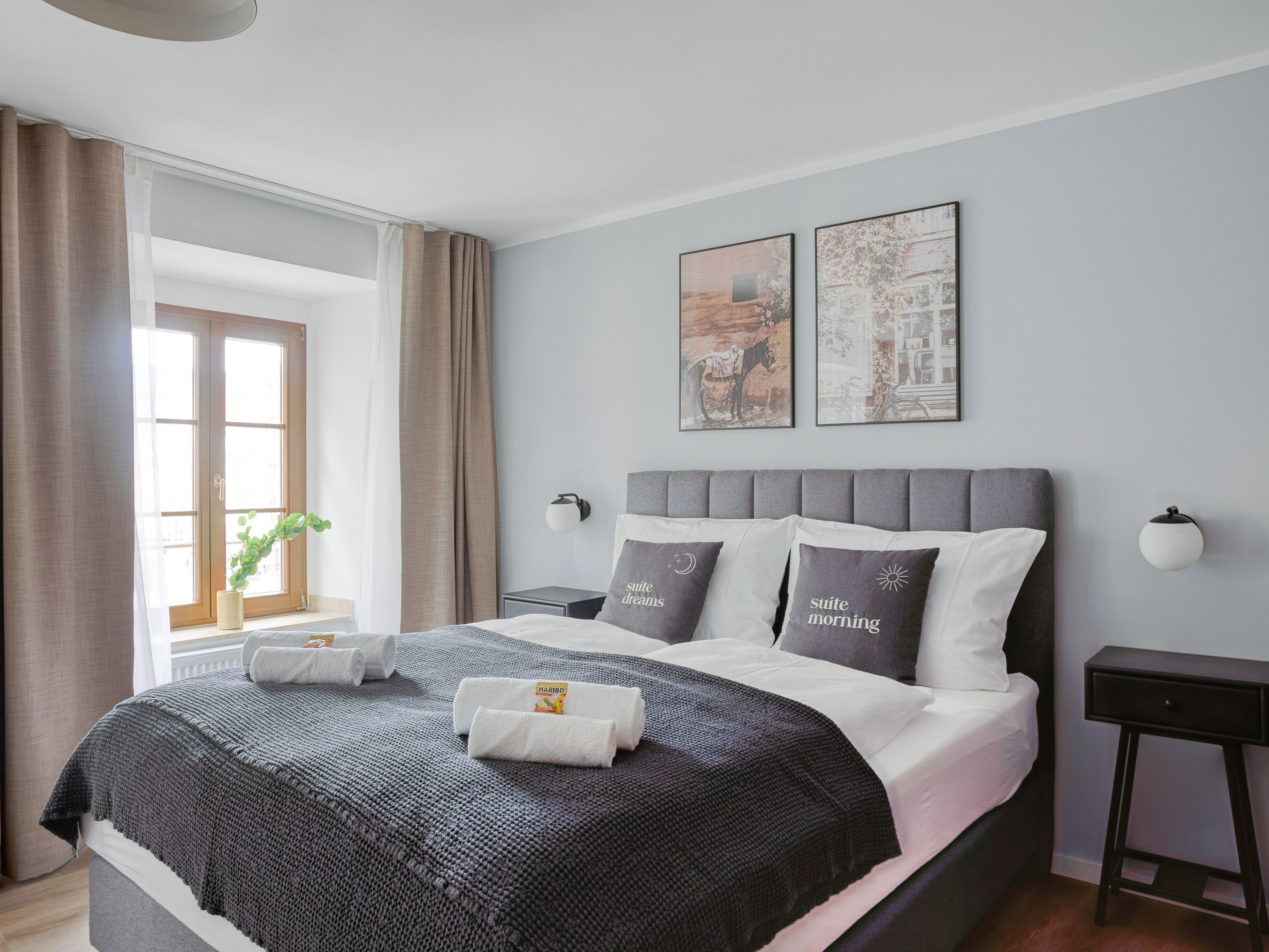 Une chambre à coucher moderne avec un grand lit, une literie aux tons de gris et de blanc, deux lampes fixées au mur et deux tableaux encadrés.