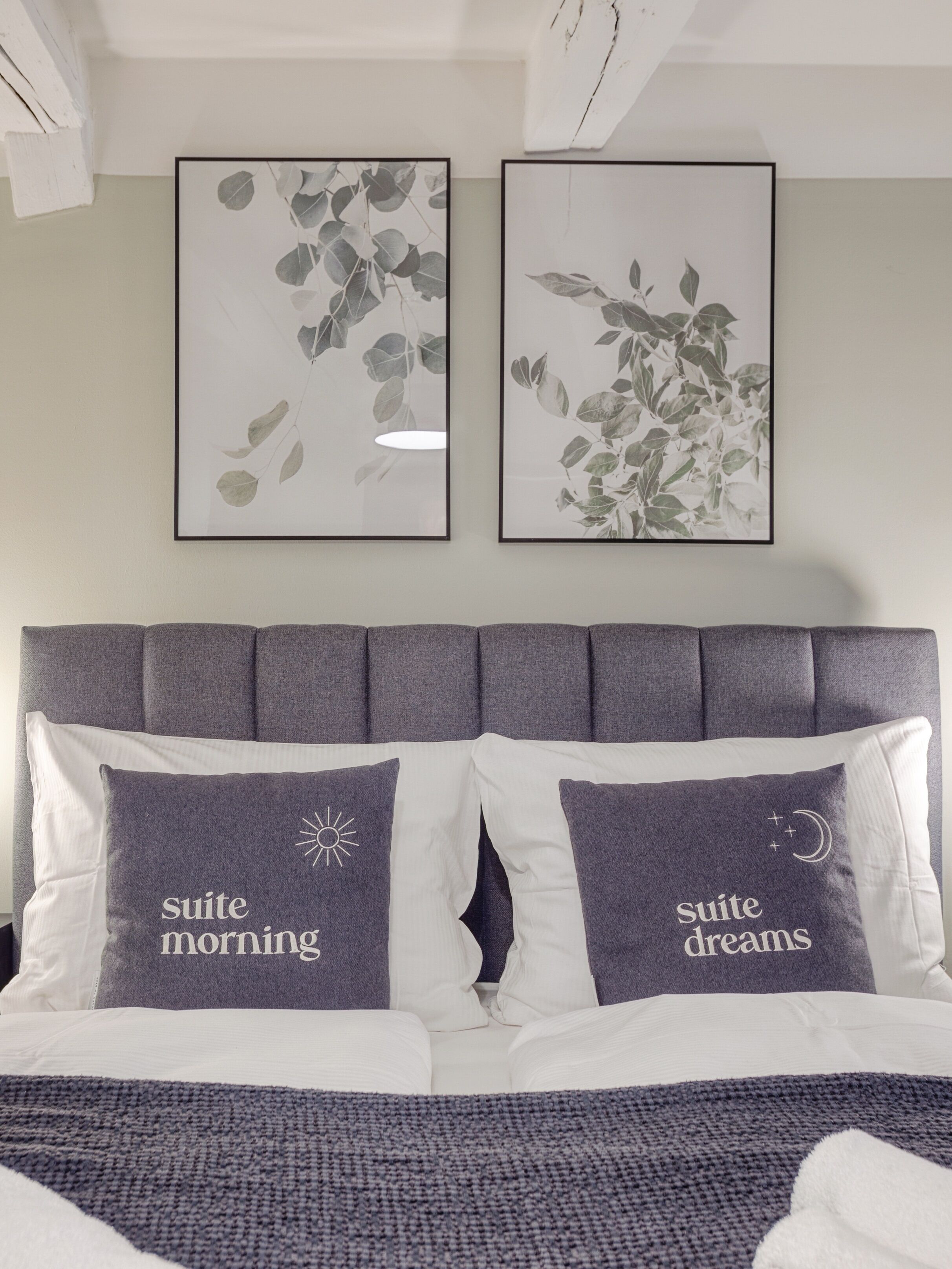 Ein Schlafzimmer mit einem grauen, gepolsterten Kopfteil, zwei Kissen mit Schriftzug und zwei gerahmten Pflanzenabbildungen an der Wand.