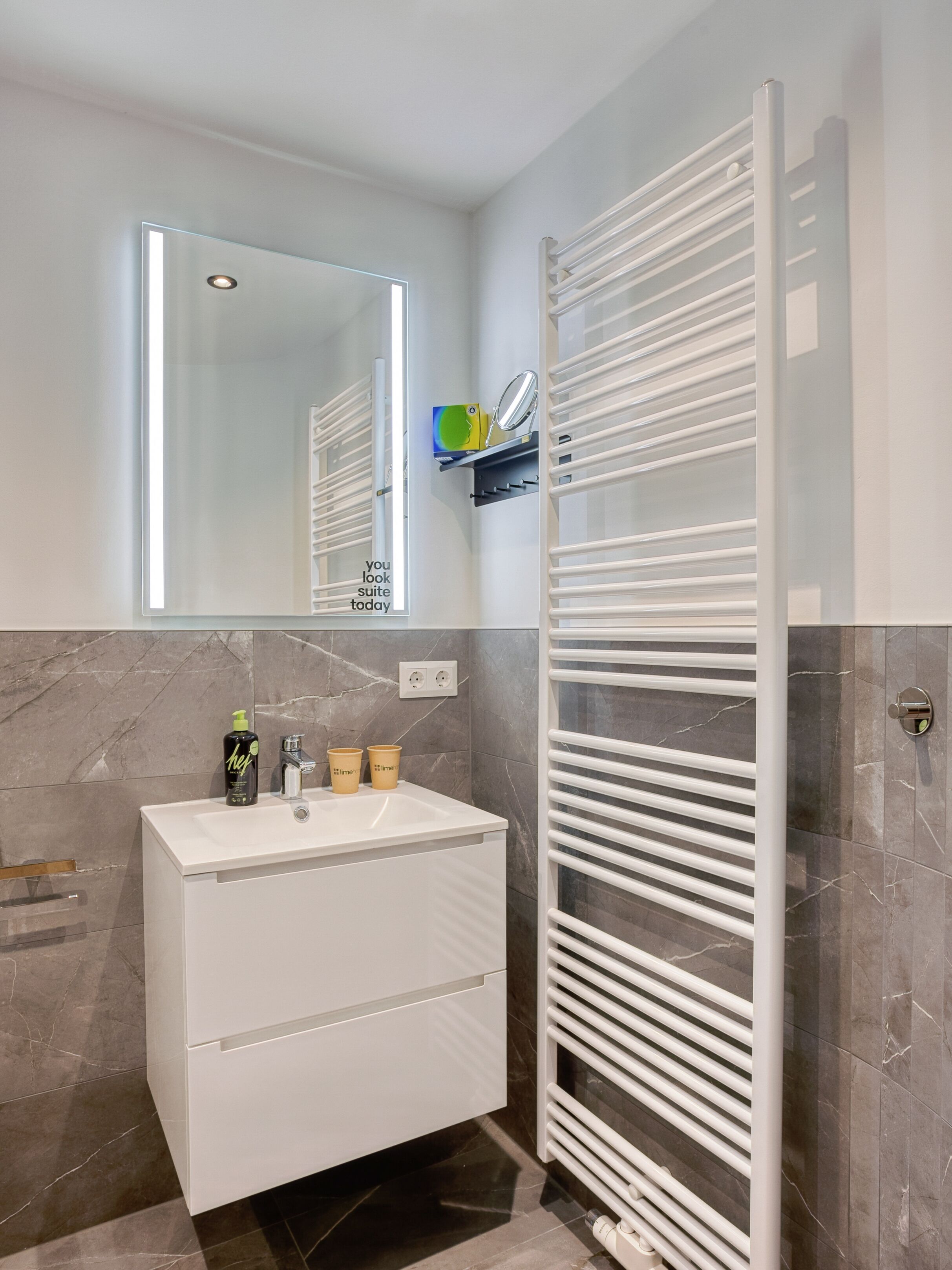 Ein modernes Badezimmer mit einem weißen Waschtisch, einem Spiegel mit integrierter Beleuchtung, einem Handtuchhalter und einer Wand mit Marmorfliesen.