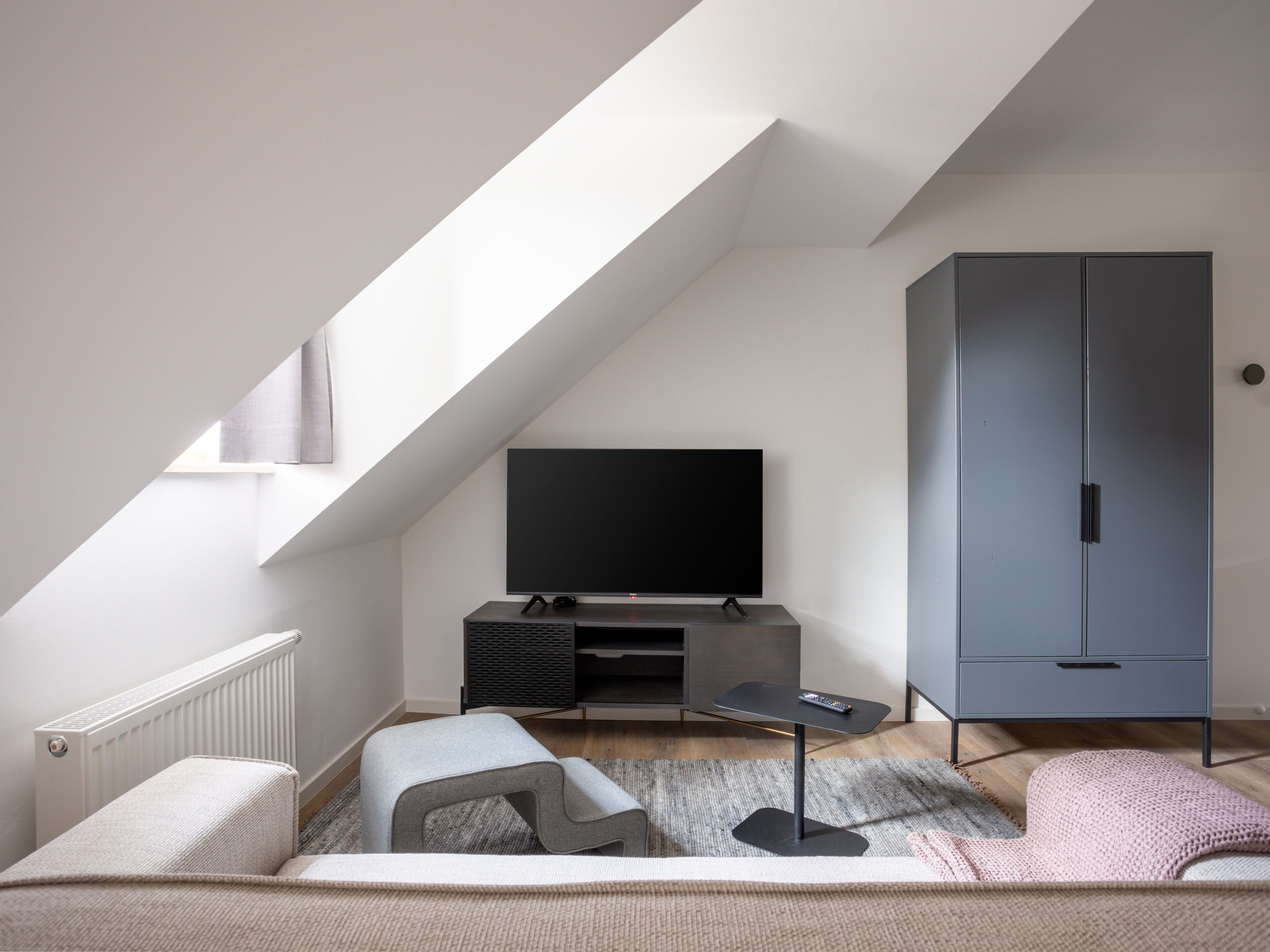 A modern loft bedroom with a sloped ceiling, a large flat-screen TV, a grey wardrobe, and a cozy seating area with a rug and cushions.