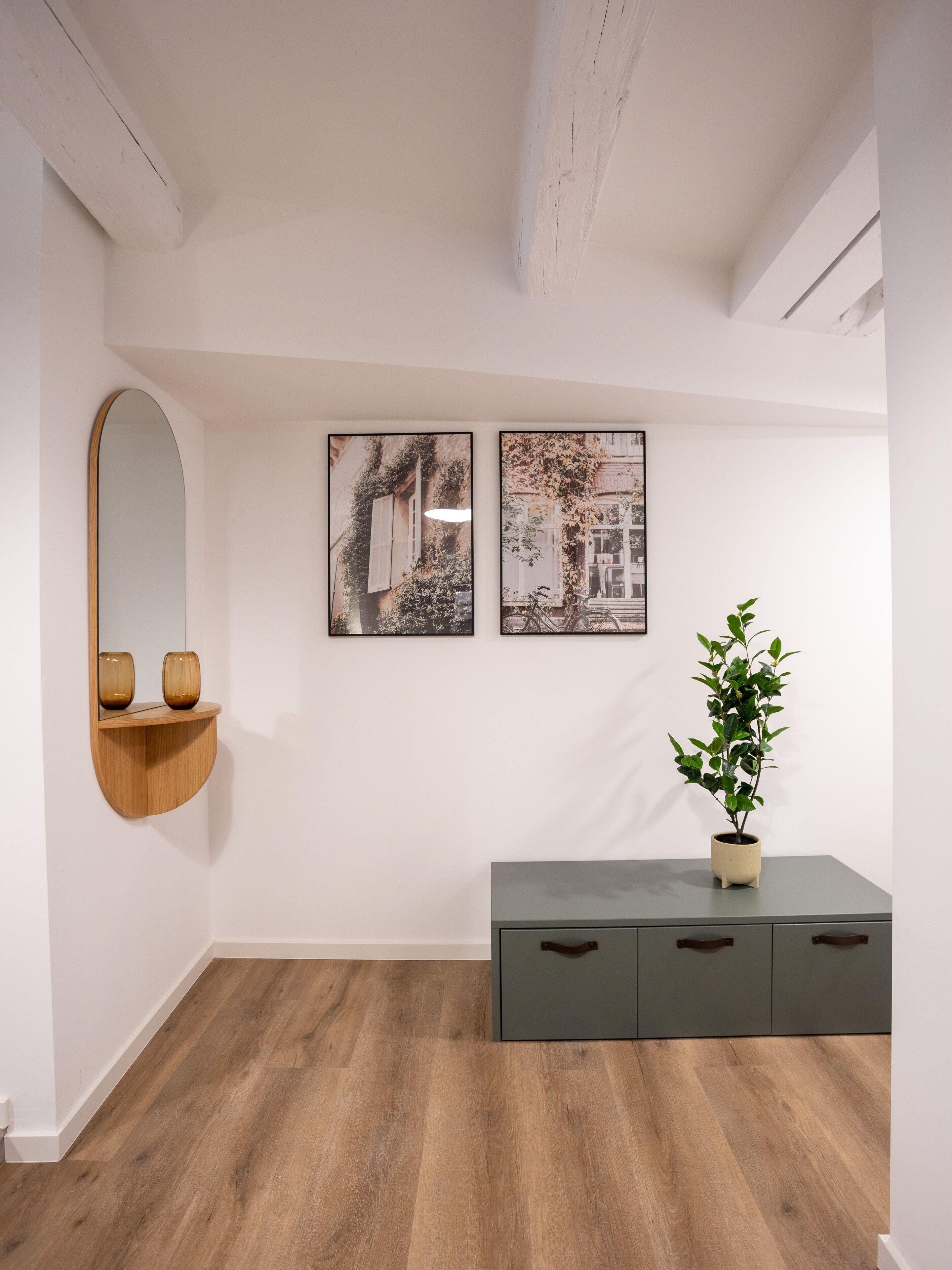 A room with a wooden floor, a grey cabinet with drawers, a mirror with a wooden frame, and two framed pictures on the wall.