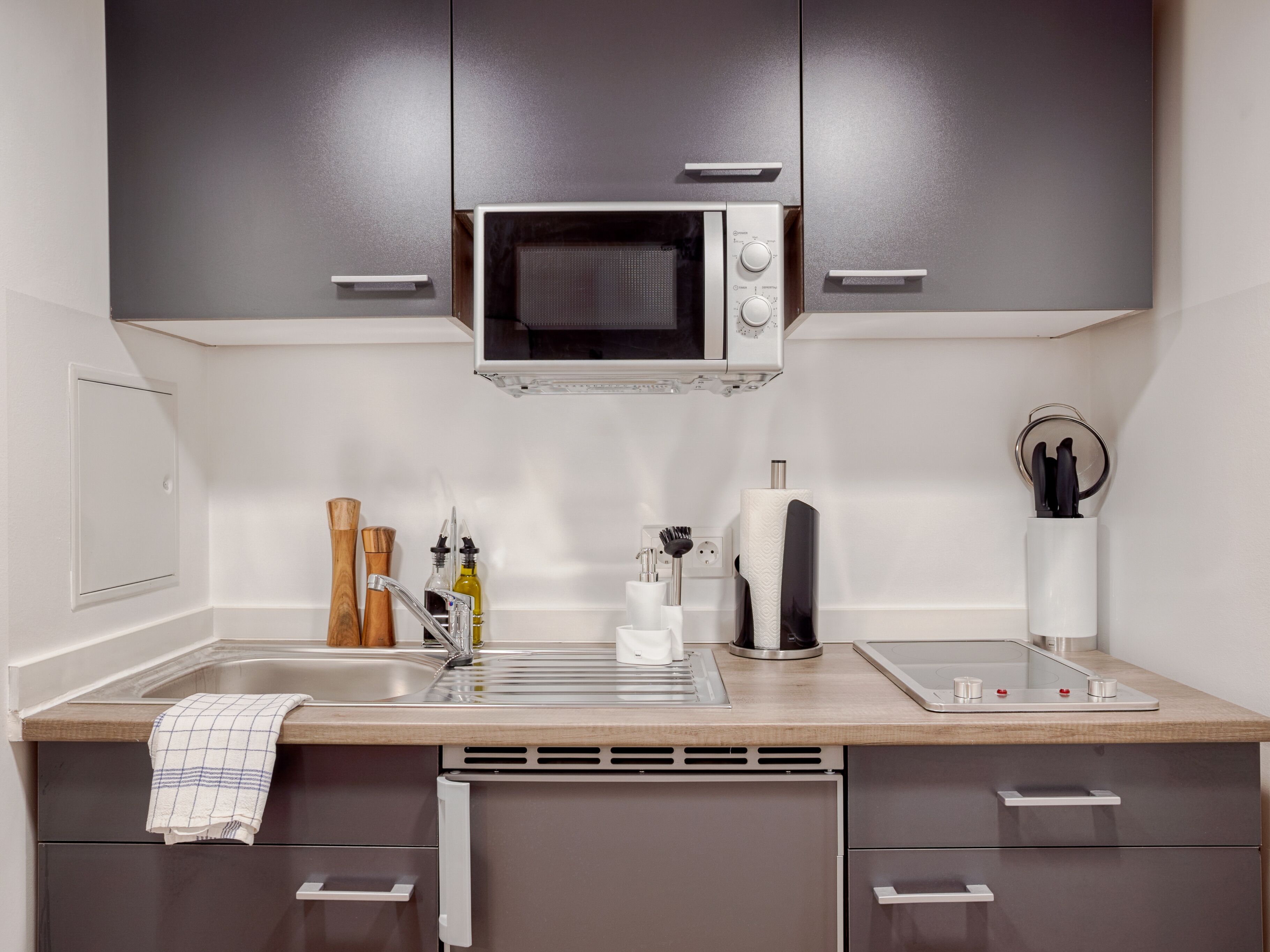 A modern kitchen with grey cabinets, a built-in microwave, a sink with a towel, and a coffee machine.