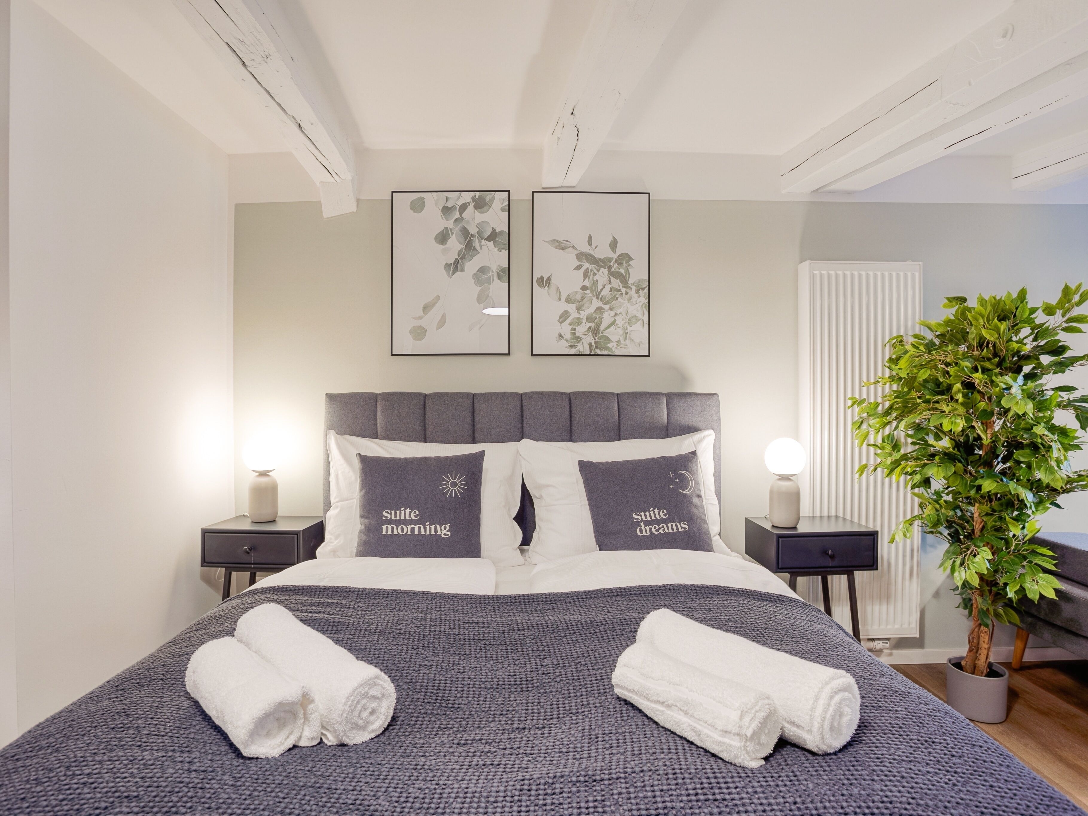 En pænt redt seng med to puder, et natbord med lampe og en potteplante.