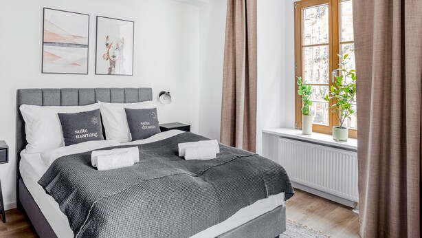 Une chambre à coucher moderne avec un lit recouvert d’un tissu gris, deux lampes fixées au mur et une fenêtre avec des rideaux.