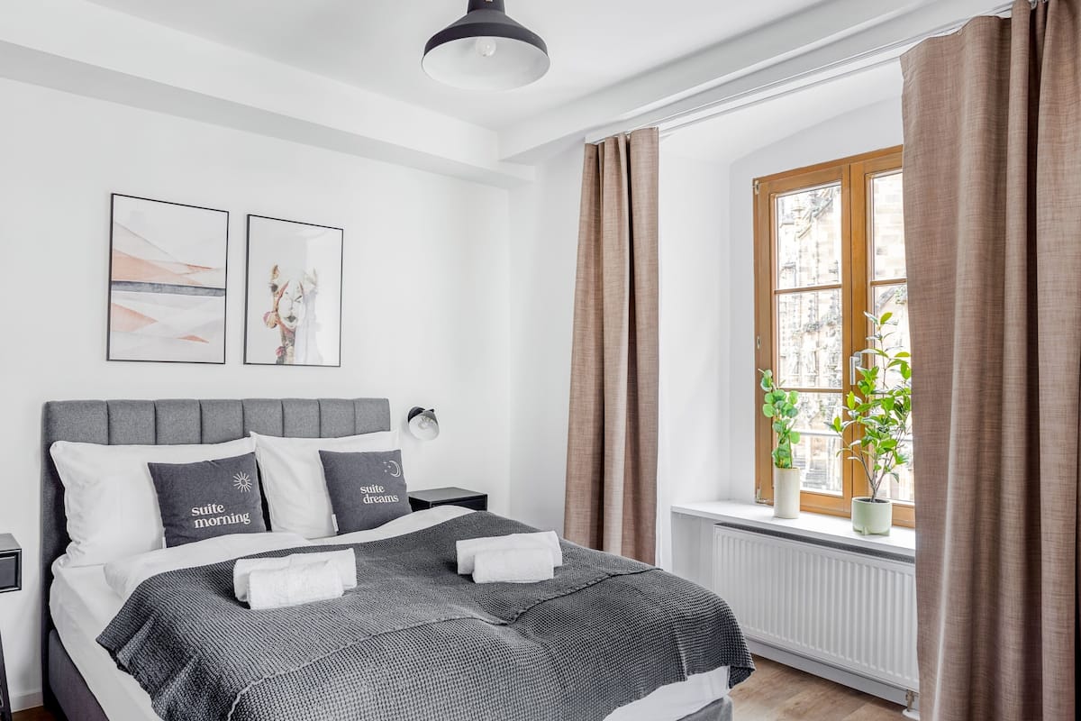 Une chambre à coucher moderne avec un lit recouvert d’un tissu gris, deux lampes fixées au mur et une fenêtre avec des rideaux.