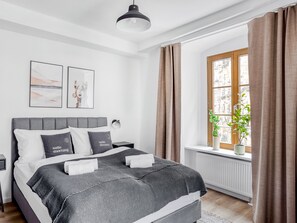 Une chambre à coucher moderne avec un lit recouvert d’un tissu gris, deux lampes fixées au mur et une fenêtre avec des rideaux.