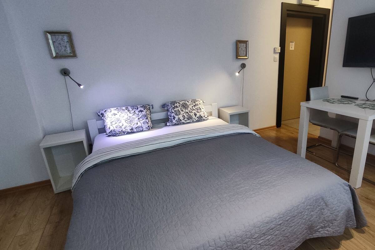 A bedroom with a bed, bedside tables, a desk with a chair, a television, and a door.