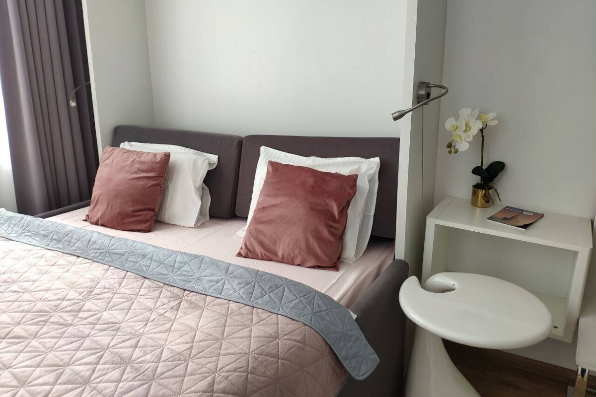 A bedroom with a bed, pillows, a bedside table with a book, and a wall-mounted shelf with a vase.
