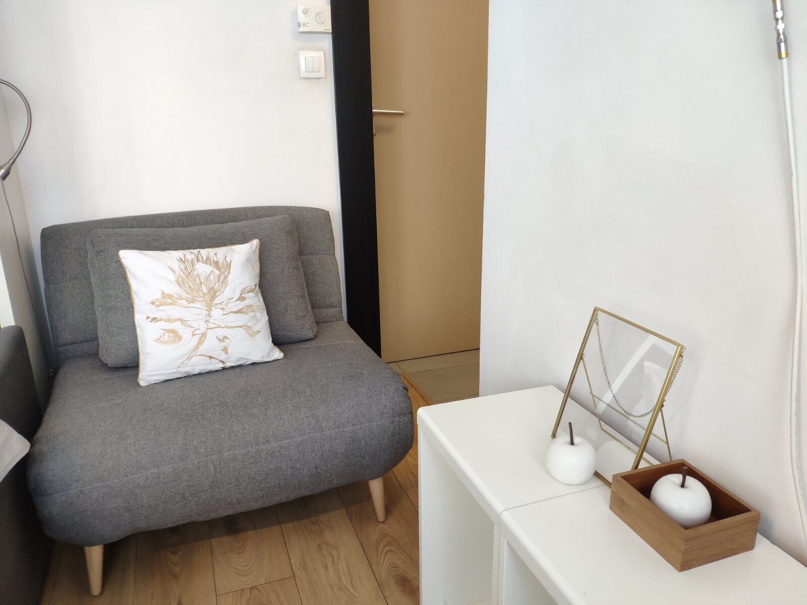 Un sofá gris con un cojín decorativo, una mesita de noche blanca con un macetero de madera en forma de manzana, y un cuadro enmarcado apoyado contra la pared.