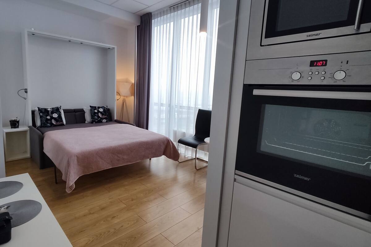 A modern hotel room with a bed, a desk with a computer, a flat-screen TV, and a built-in oven.