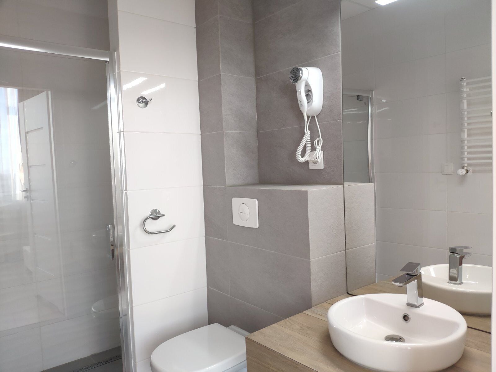 Un baño moderno con cabina de ducha de vidrio, un inodoro blanco, un secador de pelo montado en la pared y dos lavamanos con accesorios cromados.