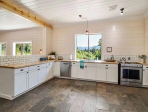 Une cuisine moderne avec des meubles blancs, un plafond en bois, une grande fenêtre donnant sur un paysage verdoyant et un four en acier inoxydable.