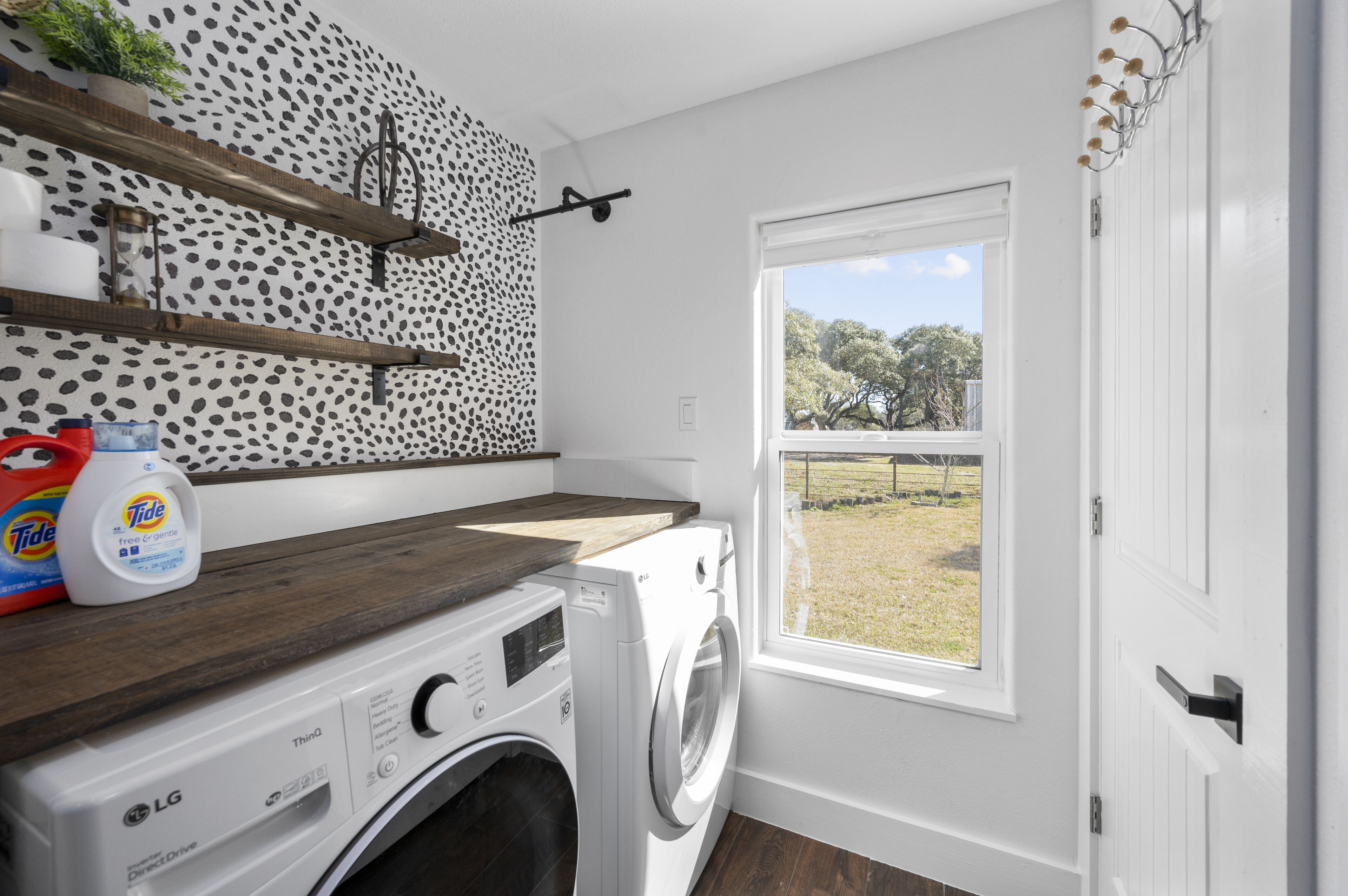 A laundry room with a washing machine, a shelf with laundry products, a window with a view of trees, and a patterned wall.