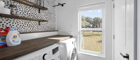 A laundry room with a washing machine, a shelf with laundry products, a window with a view of trees, and a patterned wall.