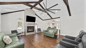 Un salon spacieux doté d’une cheminée, de canapés verts et gris, de parquet et de poutres apparentes.