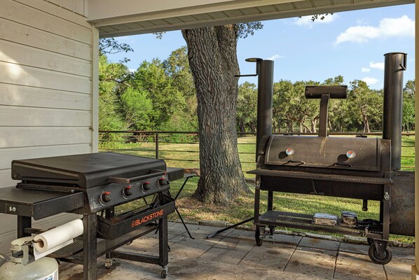 An outdoor grill setup with a gas tank, a charcoal grill, and a smoker, positioned under a covered area with a tree nearby.
