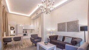 Una sala moderna con un sofá, sillones, una mesita de centro y un comedor con una mesa y sillas. Se ve una cocina al fondo con armarios blancos y una encimera de mármol.