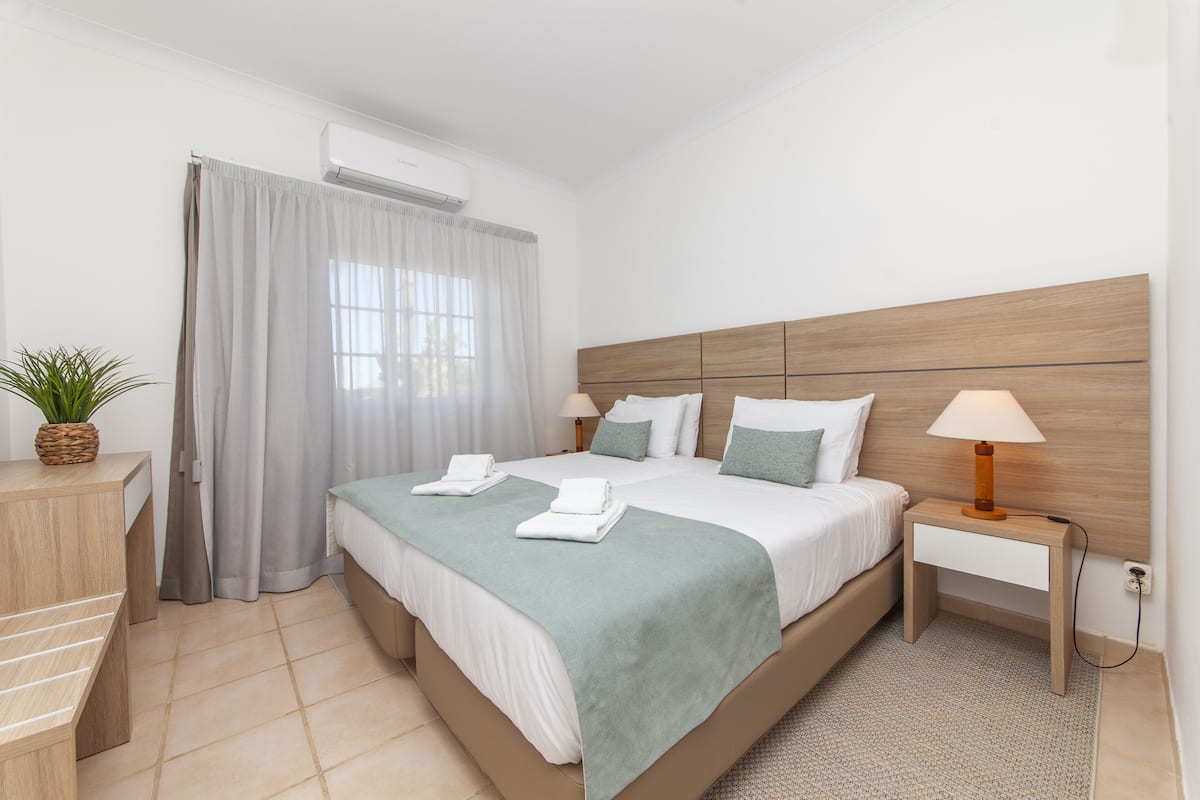 A modern hotel room with a large bed, a bedside table with a lamp, a small desk with a plant, and a window with curtains.