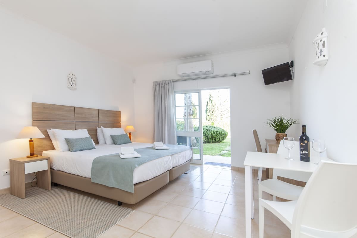 A modern bedroom with a large bed, bedside tables, a desk with chairs, a TV, and a view of the outdoors.