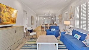 A spacious living room with a large television, blue sofas, a wooden coffee table, wicker chairs, and a dining area in the background. - Escape to The Moorings #42 (Jekyll Island)
