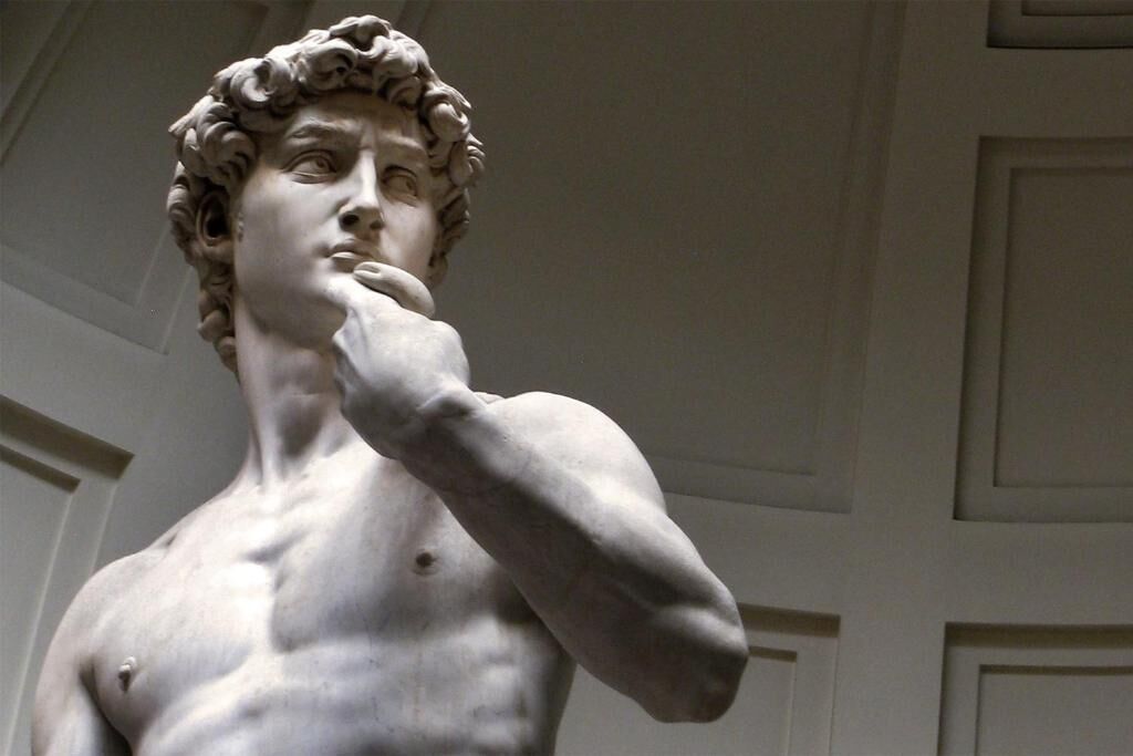 Eine aus Marmor gefertigte Statue mit muskulösem Körperbau und Locken, vor einer schlichten Wand mit geometrischem Muster.