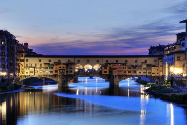Un puente histórico sobre un río tranquilo al anochecer, rodeado de edificios con ventanas iluminadas.