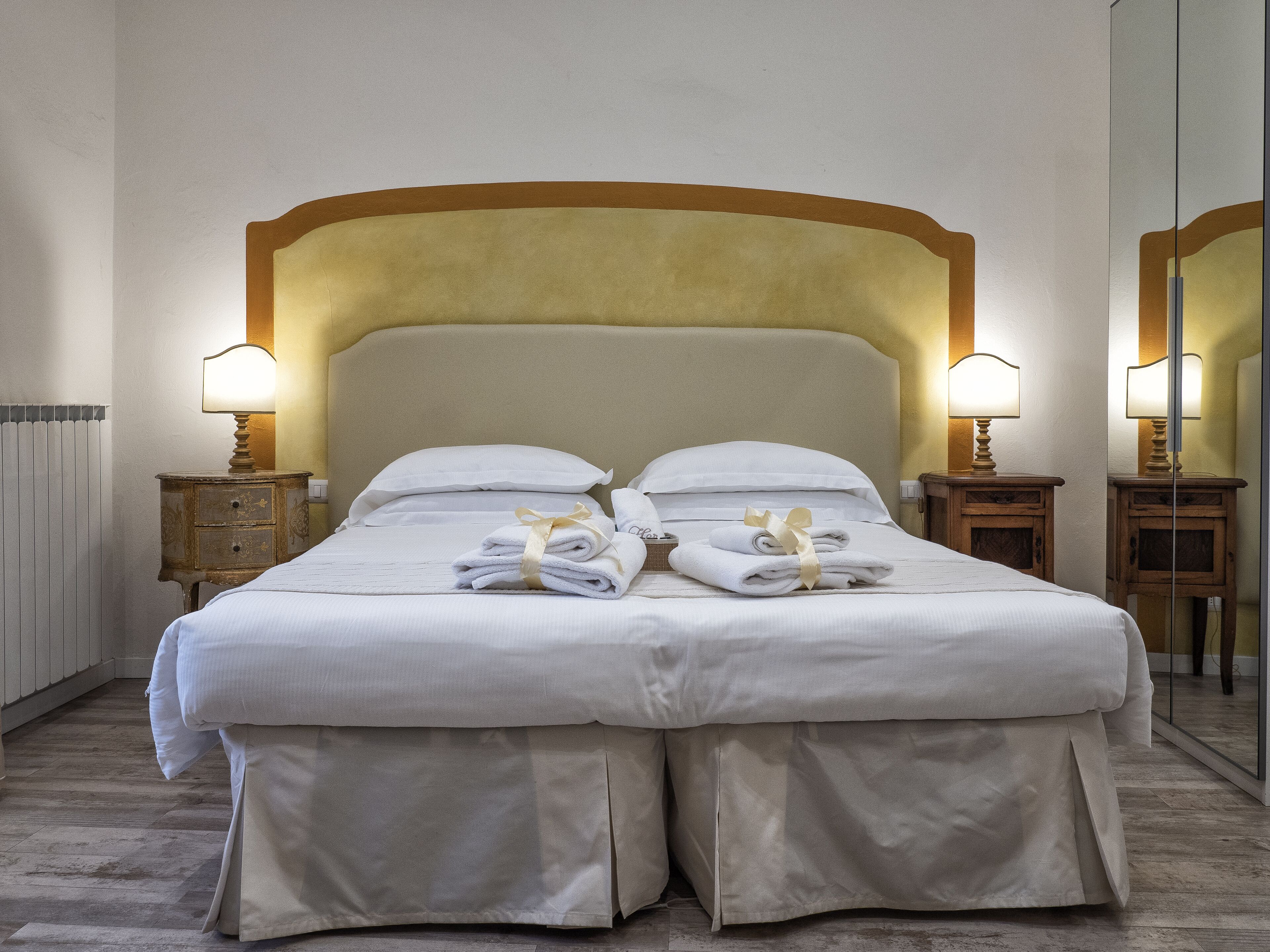 Uma cama de casal bem arrumada com lençóis brancos, duas mesas de cabeceira com abajures e um espelho grande na parede.