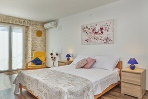 Une chambre à coucher comprenant un lit, une chaise, une table de chevet, une lampe, une œuvre d’art murale et une fenêtre donnant sur un paysage.