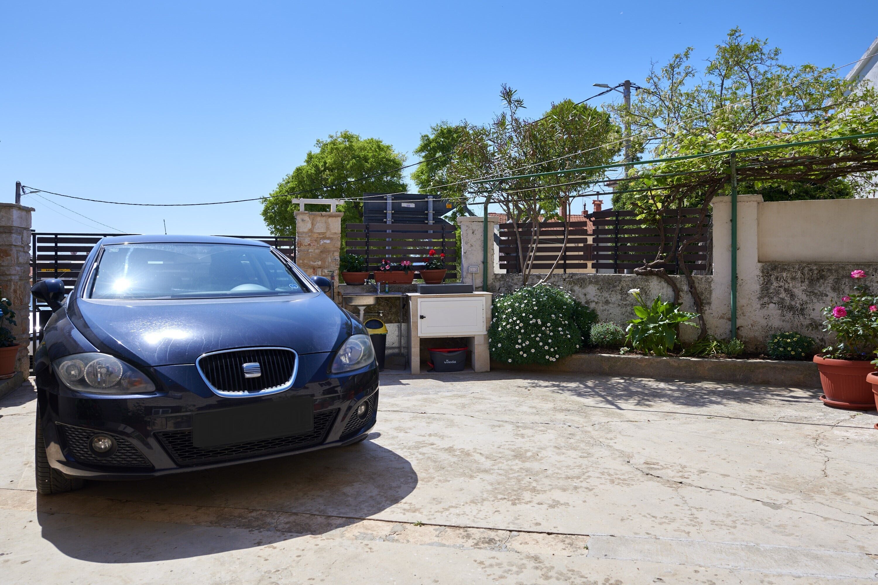 A blue car parked in a driveway with a gate, potted plants, and a building in the background.