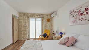 A bedroom with a stone wall, a wooden door, a bed with white and pink pillows, a yellow armchair, and a painting on the wall. - Nice apartment for 2 people, 3 stars, 5 minutes to the beach (Ližnjan)