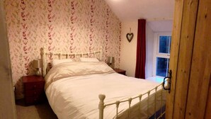 A bedroom with a white metal bed frame, floral wallpaper, a wooden door, and a window with a red curtain. - Yr Hen Siop (Betws-Y-Coed)