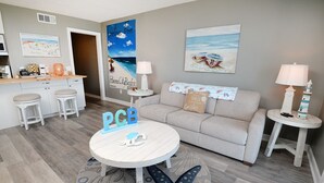 A modern living room with a grey sofa, a round coffee table, and a kitchen area with a white countertop and bar stools. - Beachside Escape: Pool, Balcony, Free Chairs & More (Panama City Beach)
