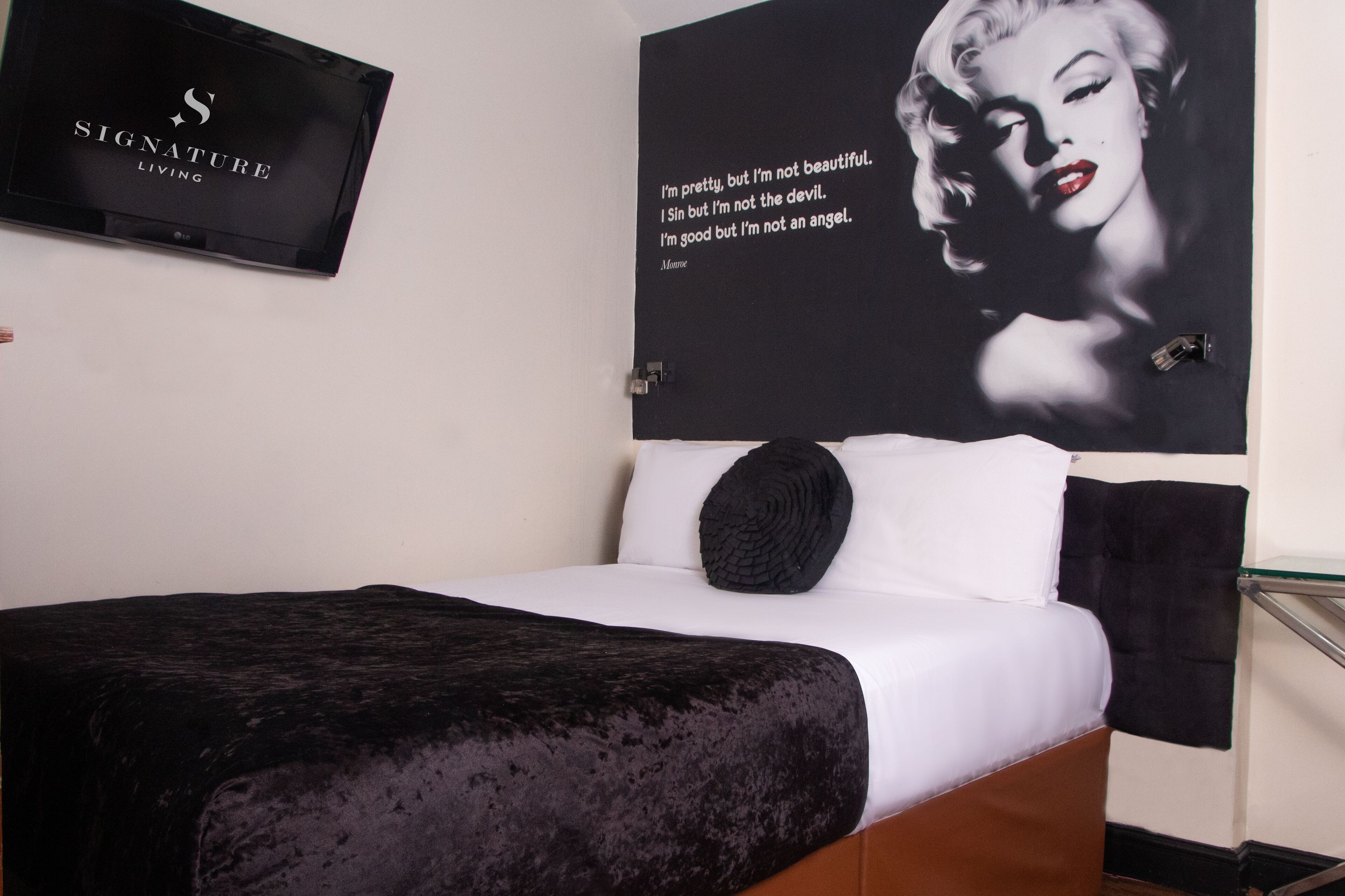 A hotel room with a bed, a black throw blanket, a black pillow, a black wall art with text, and a black sign on the wall.