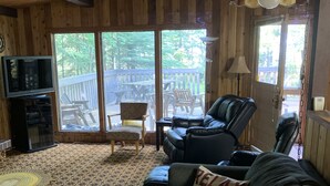 L’intérieur chaleureux d’un chalet avec un plafond en bois, une grande fenêtre donnant sur une terrasse, un canapé en cuir, un tapis à motifs et un téléviseur posé sur un meuble.