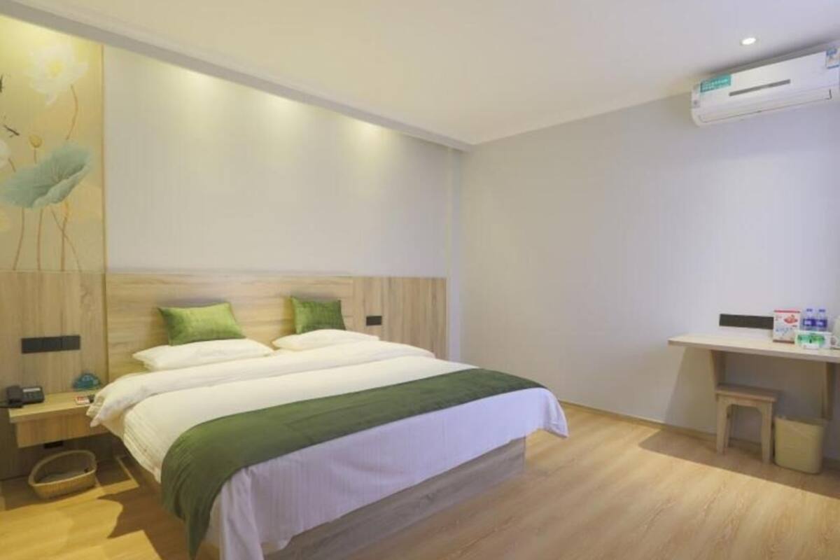 A hotel room with a bed, bedside table, nightstand, wall-mounted air conditioning unit, and a small wooden stool.