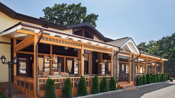 A wooden outdoor seating area with tables and chairs, a covered walkway, and neatly trimmed hedges.