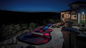 A house with a large guitar-shaped pool illuminated at night, surrounded by a landscaped garden and a hilly landscape. - Austin Guitar Pool | 87.5 FT Guitar Pool (Austin)
