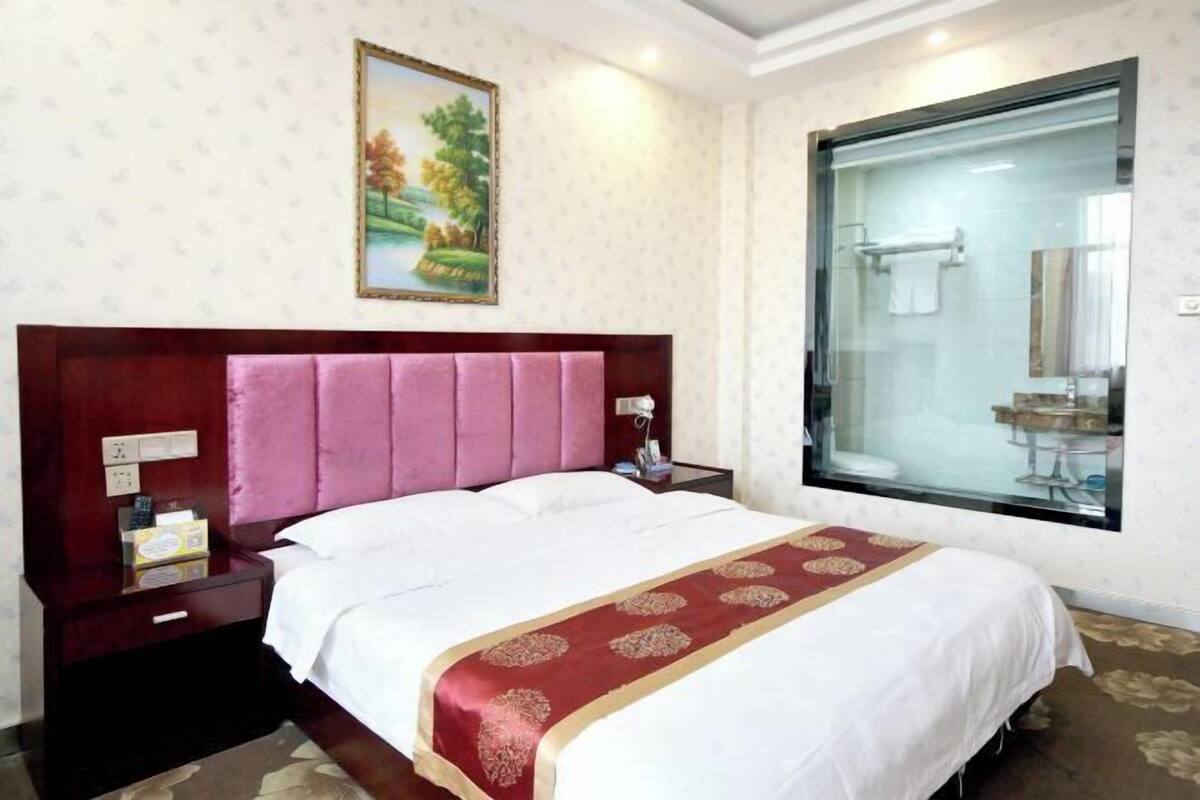 Habitación de hotel con una cama grande, una mesita de noche con lámpara, un cuadro en la pared y un ventanal con vistas.