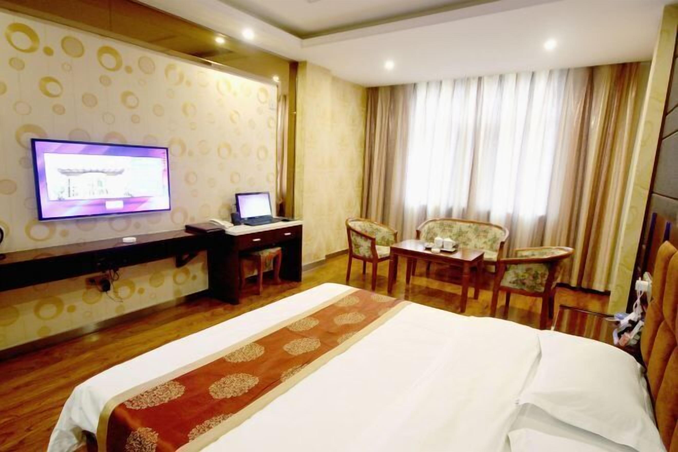 A hotel room with a bed, a desk with a laptop, a television, and a seating area with chairs.