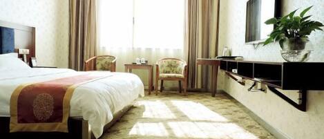 A hotel room with a large bed, a TV mounted on the wall, a small table, a chair, and a potted plant.