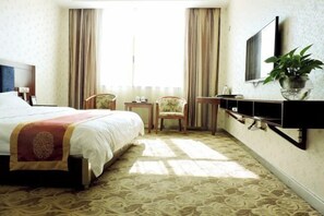 Ein Hotelzimmer mit einem großen Bett, einem an der Wand montierten Fernseher, einem kleinen Tisch, einem Sessel und einer Zimmerpflanze.