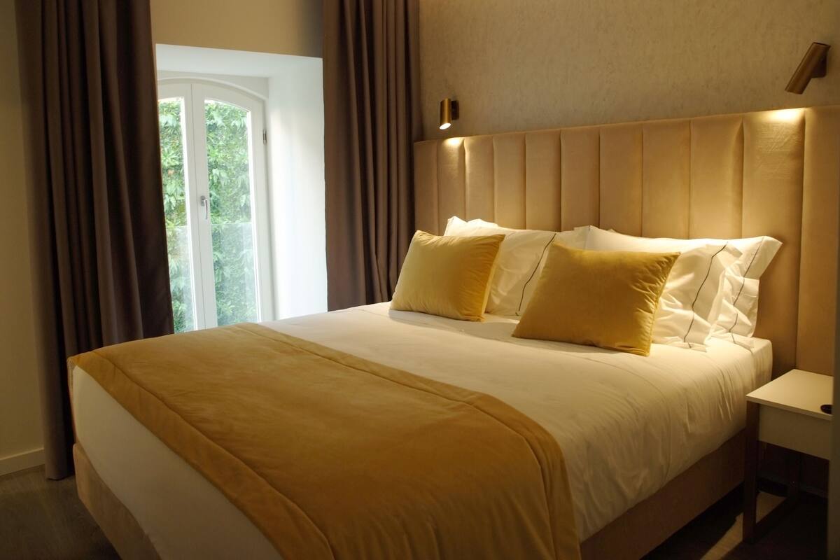 A large bed with a headboard, a bedside table, and a window with curtains.
