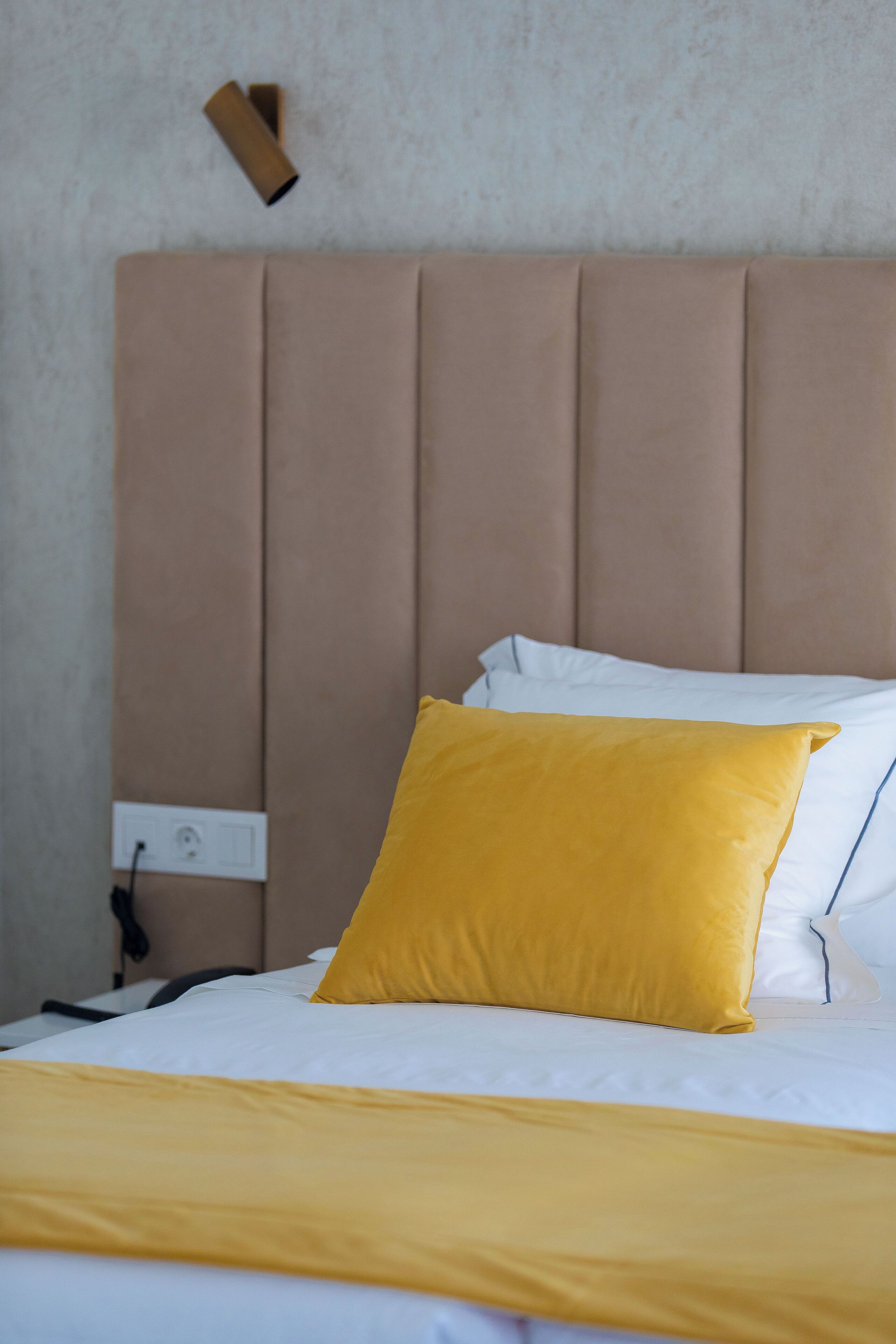 A bed with a beige headboard, white bedding, and a yellow accent pillow.