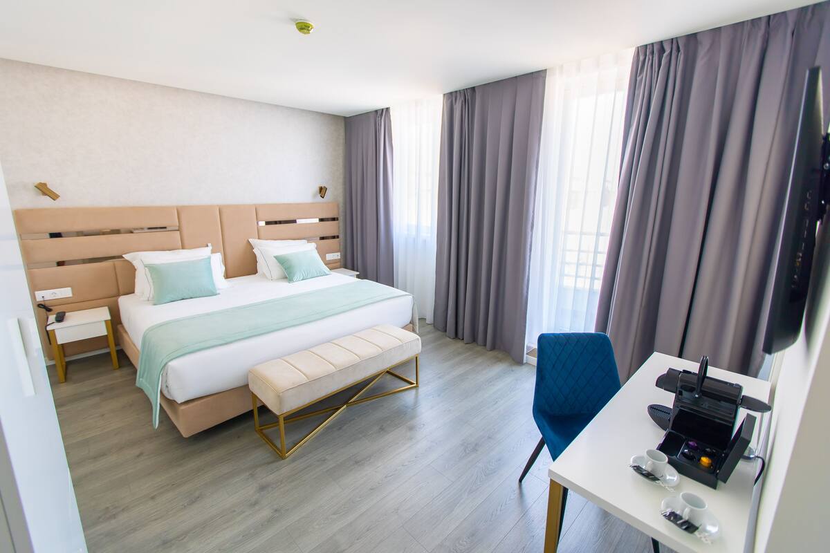 A modern hotel room with a large bed, a bench, a desk with a coffee maker, and a TV.